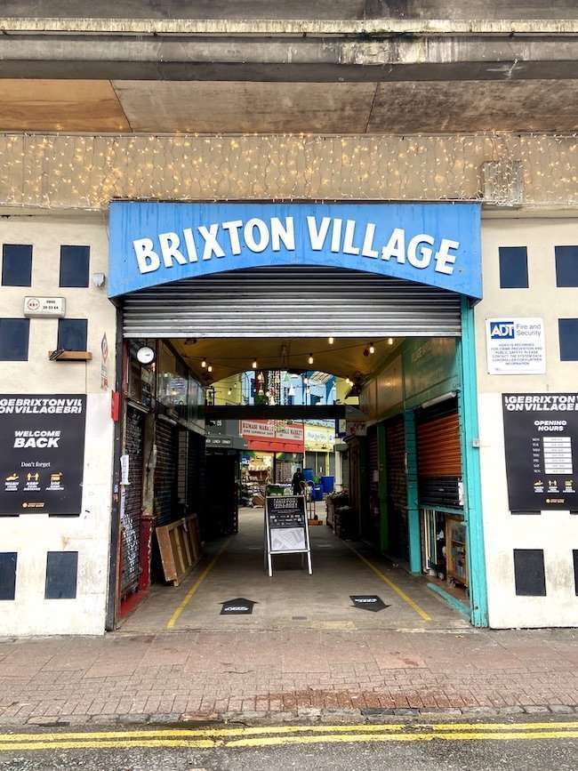 Bustling interior arcade of Brixton Village filled with independent food stalls, restaurants, and lively diners under a historic glass roof.